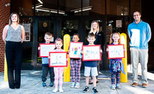 Five local elementary school children lined up beside three teachers with their Safety Week drawings for the safety calendar drawing contest.