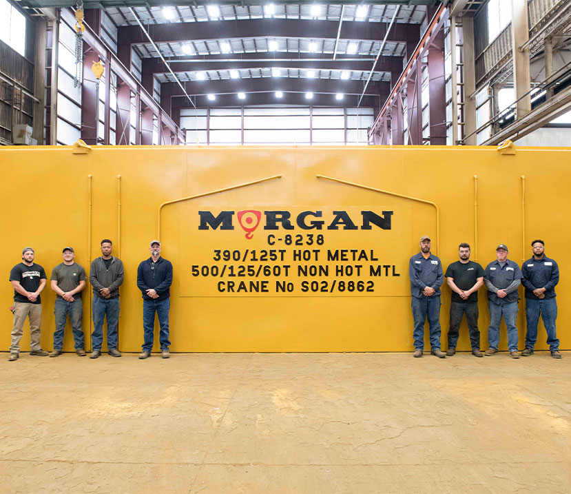 Eight men standing in front of a newly assembled yellow Morgan Crane in the Morgan warehouse.